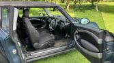 A blue mini car with the driver’s door open reveals a black and grey interior. Parked on grass near an orchard of green trees, the steering wheel, dashboard, and front seat are clearly visible as sunlight brightens the scene.