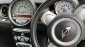 Close-up view of a MINI car dashboard and steering wheel. The large central speedometer displays mph, with a smaller tachometer behind the wheel. Air vents, controls, and MINI logo highlight the distinctive style loved by any MINI trader.