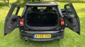 A black Mini Clubman car with both rear barn doors and side doors open, revealing the empty trunk and rear seats, is parked on green grass amid trees and bushes. The UK license plate AY59 ZSE is visible—perfect for any Mini trader.