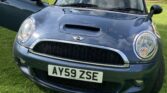 A blue Mini car with its driver’s side door open is parked on a green lawn. The front license plate reads “AY59 ZSE.” Trees and bushes surround the mini, set against a partly cloudy sky.