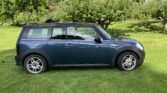 A dark blue Mini Clubman with a black roof is parked on green grass in front of leafy trees. The stylish mini car is seen from the side, showing two doors and five-spoke alloy wheels, set in a sunny garden with dappled sunlight.