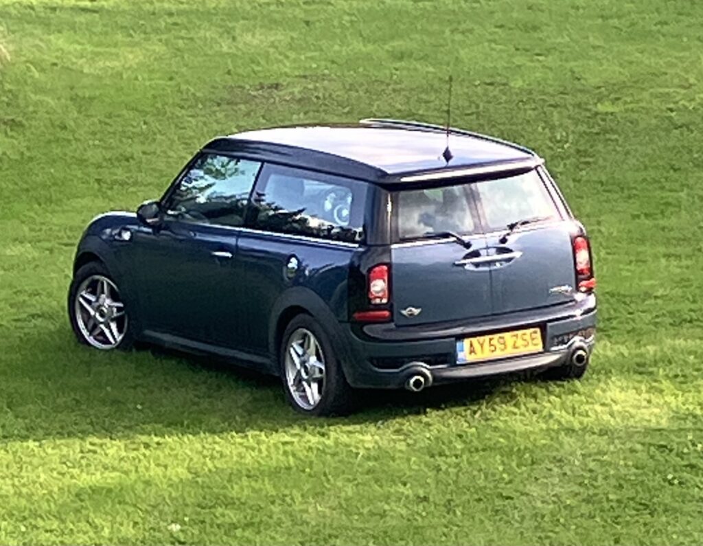 A dark blue Mini Clubman with silver trim is parked on green grass. This stylish mini car features two-part split rear doors and a yellow license plate reading "AY59 ZSE." The scene is outdoors, under daylight.