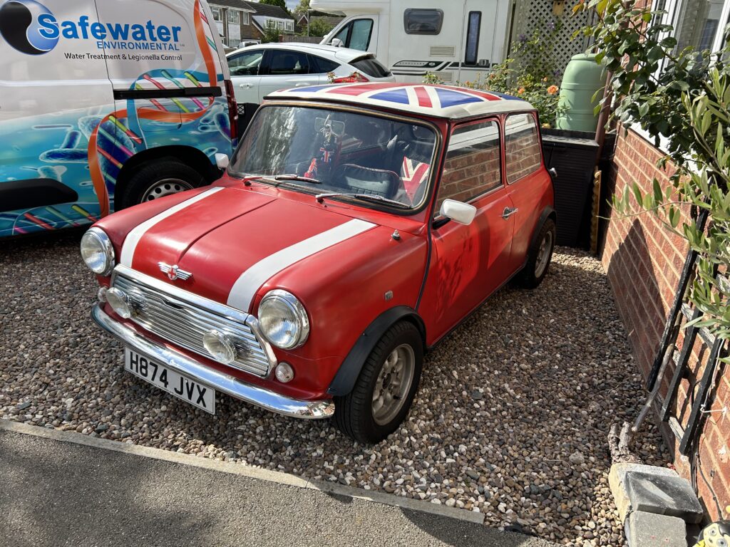 A classic red mini car with white racing stripes and a Union Jack roof is parked on a gravel driveway next to a brick house. The license plate reads "H874 JVX." A white van with “Safewater ENVIRONMENTAL” is nearby, perfect for any mini trader's eye.