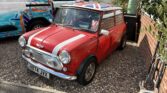 A classic red mini car with white racing stripes and a Union Jack roof is parked on a gravel driveway next to a brick house. The license plate reads "H874 JVX." A white van with “Safewater ENVIRONMENTAL” is nearby, perfect for any mini trader's eye.