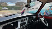 Interior of a classic mini car with a black-and-silver dashboard. A British flag boxing glove and fuzzy dice hang from the mirror. Through the window, houses, a van with "mini trader" text, and a rainbow steering wheel cover are visible outside.