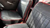 Close-up of the front seats in a classic mini car. The seats are black with red piping and vertical stitching. The door is black with a silver manual window crank, and a Union Jack pillow rests on the rear seat—a true find for any mini trader.