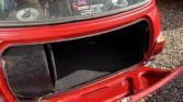 Open trunk of a red classic mini car with no license plate attached. A sticker on the rear window reads, “SORRY OFFICER, THOUGHT YOU WANTED A RACE” with a small British flag above it. Reflections and gravel surround the mini.