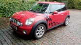 A red mini car with a black-and-white checkered flag decal on the side is parked on a wet cobblestone road, surrounded by green leafy trees and bushes. The mini has silver wheels and a white roof.