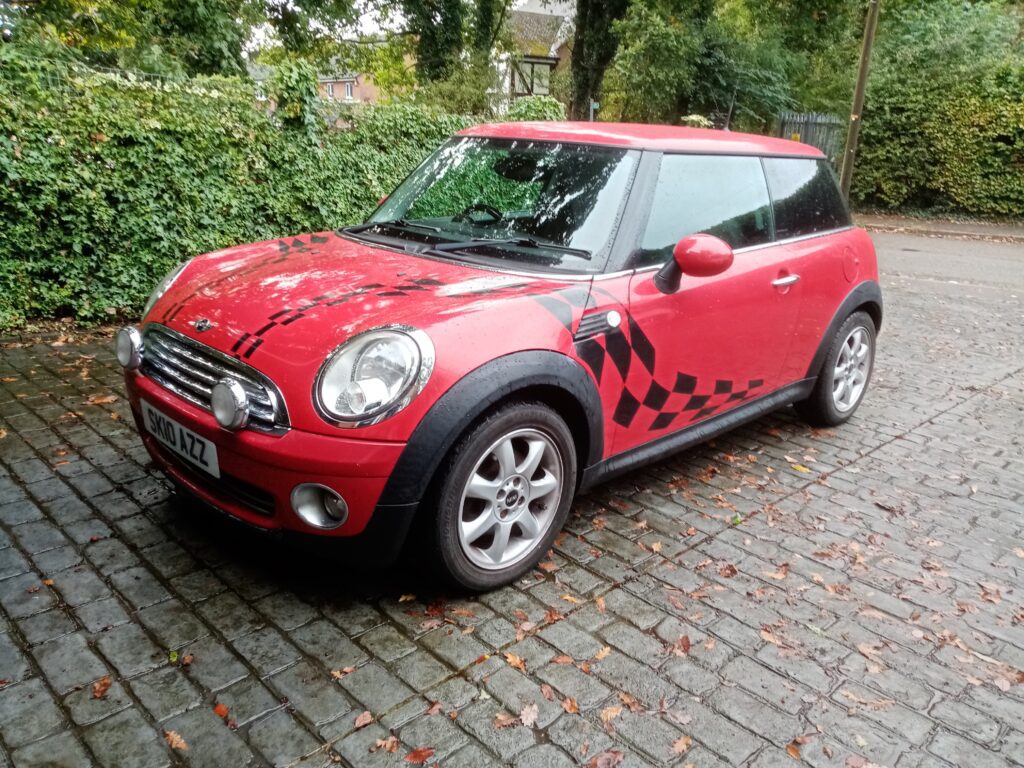 A red mini car with black checkered decal on the side is parked on a wet cobblestone driveway. The car has silver rims, round headlights, and extra front lights. Green hedges and trees are in the background on a cloudy day.