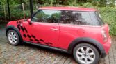 A red Mini Cooper from Mini Trader with black checkered flag decals is parked on a cobblestone driveway. The mini car features silver alloy wheels and black-tinted windows, surrounded by leafy green bushes and trees in the background.