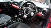 Interior of a mini car showing the steering wheel, dashboard with a large central speedometer, manual gear stick, black seats with gray fabric, and red trim accents. This stylish Mini is parked outside near a brick house—perfect for any mini trader.