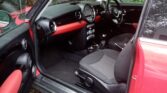 A red mini car with its driver’s door open reveals a black and red interior, round dashboard dials, and a manual gear stick. The car is parked on stone paving among fallen leaves, with a green wooded area in the background—ideal for any mini trader.