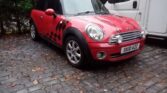 A red Mini Cooper with black racing stripes and a checkered pattern on the side is parked on a cobblestone driveway. This stylish mini car, with UK plate "SK10 AZZ," sits beside a white van amid autumn leaves. Perfect for any mini trader’s collection.