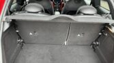 View of an open mini car trunk with rear seats upright; black carpeted cargo area is empty. Two headrests, dashboard with circular center display, and climate controls are visible. Interior features stylish black and gray tones.