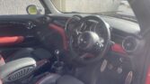 Interior of a modern mini car with black and red seats, manual gear shift, three-spoke steering wheel featuring the Mini logo, round air vents, and metallic foot pedals. Sunlight streams through the window, illuminating the dashboard—a dream for any mini trader.