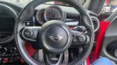 A close-up view of a Mini car's black steering wheel with the Mini logo in the center, dashboard controls behind it, and speedometer visible. The car’s interior is mostly black with red accents, and a person’s blue-clad leg is partially visible on the right.
