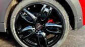 Close-up of a shiny black alloy wheel with five spokes on a red mini car. The tire is clean, showing tread detail. The red brake caliper is visible behind the wheel. The car’s black fender and wet asphalt complete this classic mini look.