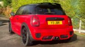A bright red mini car with a black roof and black wheels is parked on a driveway. The car’s rear and license plate, "Y3 JBR," are visible, while green trees and bushes line the driveway in the sunlight.