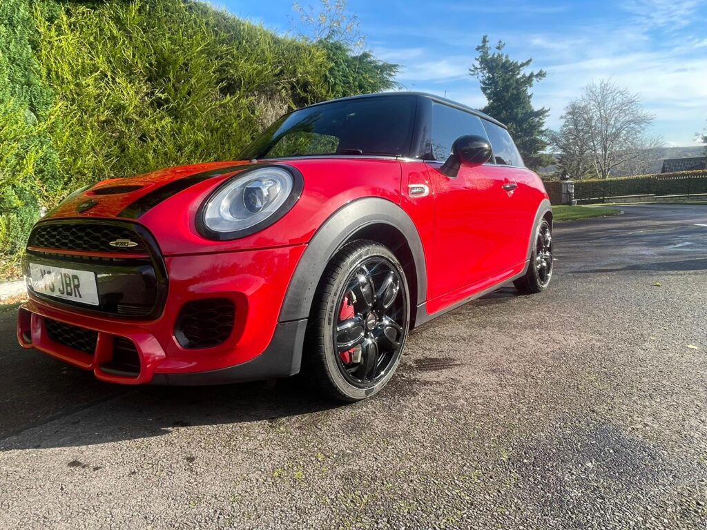 A shiny red Mini Cooper with black roof and wheels is parked on a sunlit driveway next to green bushes. The sky is blue with a few clouds, trees in the background—perfect for any Mini trader seeking a standout mini car.