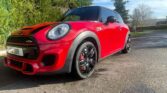 A shiny red Mini Cooper with black roof and wheels is parked on a sunlit driveway next to green bushes. The sky is blue with a few clouds, trees in the background—perfect for any Mini trader seeking a standout mini car.