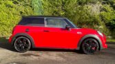 A bright red mini car with black wheels and a black roof is parked on a wet driveway. Behind the car is a dense, green hedge under sunlight, casting shadows and highlighting the shiny, clean surface—perfect for any mini trader.