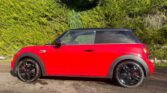 A bright red MINI car with a black roof and black alloy wheels is parked on a sunlit driveway. Dense, green hedges and tall trees form the background, casting shadows on the shiny mini and ground. The weather appears clear and sunny.