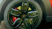 Close-up of a glossy black alloy wheel with five spokes on a mini car. The wheel features a red brake caliper behind it and is fitted with a SETULA S-RACE tire. Part of the red mini body and black wheel arch are visible.