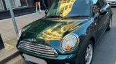 A dark green Mini car with silver accents is parked on a city street. Reflections of nearby buildings appear on its hood and windshield. Two people, perhaps mini trader enthusiasts, stand nearby facing a shop window. The license plate is blurred.