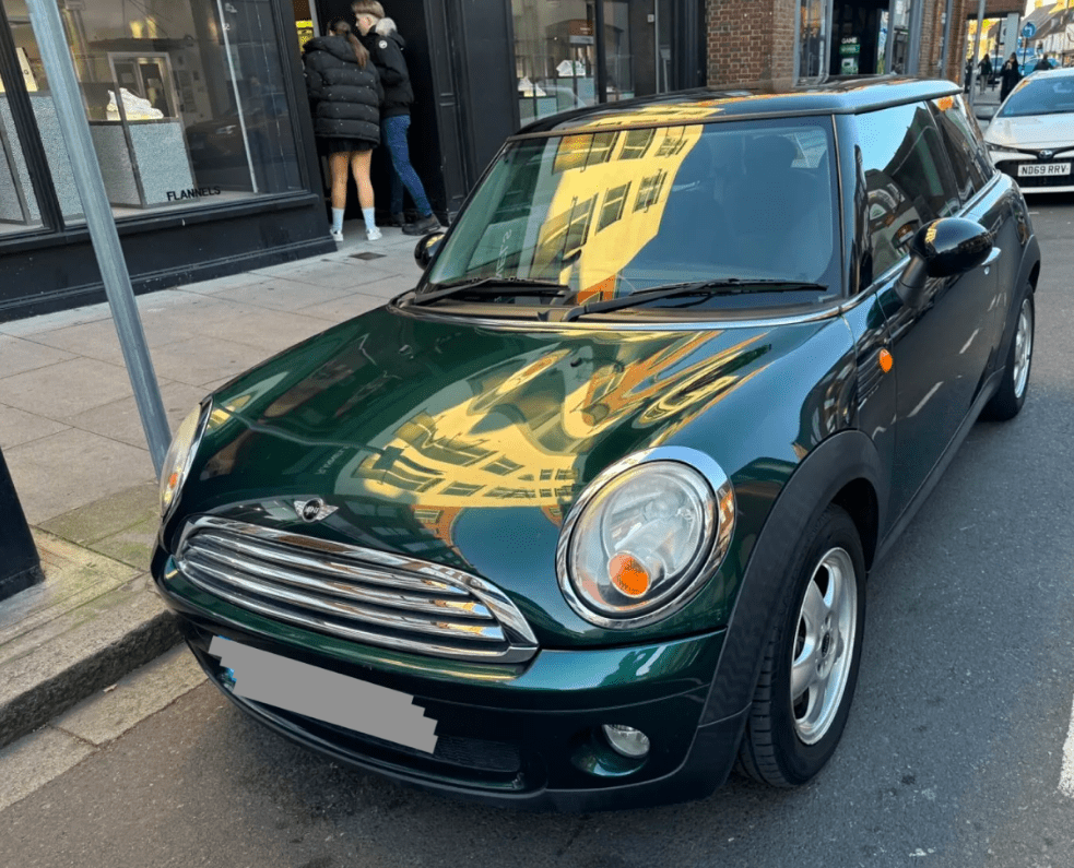 A dark green Mini car with silver accents is parked on a city street. Reflections of nearby buildings appear on its hood and windshield. Two people, perhaps mini trader enthusiasts, stand nearby facing a shop window. The license plate is blurred.