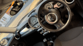 The image shows the interior of a MINI car with a right-hand steering wheel, circular air vents, a large central speedometer, black seats, and manual gear stick. Sunlight pours in through the windows, highlighting the classic MINI dashboard and controls.