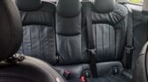 The image shows the black leather back seat of a mini car. There are three seatbacks with stitching detail, headrests, two visible seat belts, and red buckles. Outside the rear window, blurry greenery and parked cars are visible.