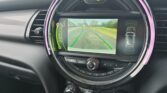 A mini car’s dashboard display shows a rearview camera image with colored guide lines. The car is in reverse, and the display includes parking sensors and climate control dials below. The interior features black surfaces and a circular center screen.