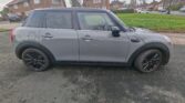 A grey Mini Cooper 5-door hatchback, ideal for any mini car enthusiast or mini trader, sits on a paved surface. Red-brick houses, grassy areas, and leafless trees are seen in the background beneath an overcast sky.