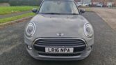 A gray Mini Cooper mini car is parked on a residential street facing the camera. The license plate reads "LR16 NPA." Brick houses, a large leafless tree, green grass, and a cloudy sky form the backdrop. Wet ground hints at recent rain.