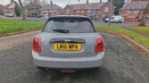 A grey Mini car with a black roof is parked on a suburban street. Viewed from the rear, its taillights and yellow plate "LR16 NPA" stand out. Brick houses, grass, trees, and other parked cars complete this classic mini trader scene.
