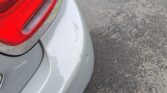 Close-up of a silver mini car's rear bumper showing scratches and cracks near the left taillight. The damage appears white against the gray paint as the car sits on asphalt with scattered brown leaves at the pavement’s edge.