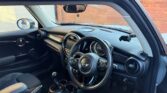 Interior view of a Mini car, featuring the black dashboard, round central display, three-spoke steering wheel with logo, manual gear stick, cup holders, and black leather seats. Sunlight filters in through the window, with a brick wall outside.