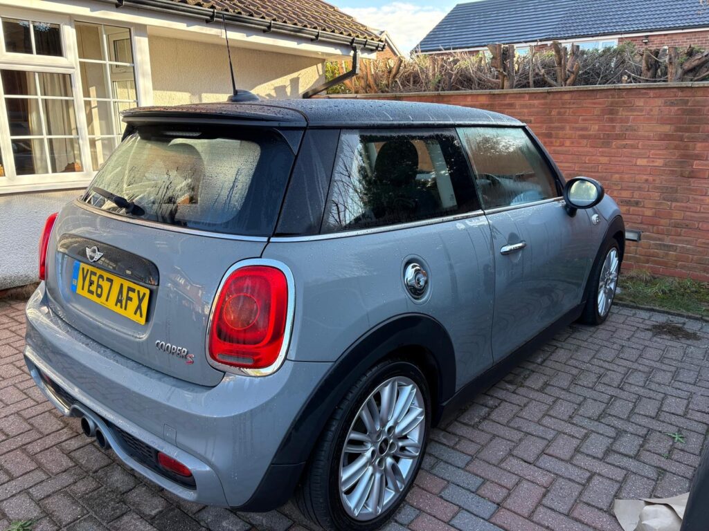 A grey Mini car is parked on a brick driveway next to a house with white-framed windows. The license plate reads "YE67 AFX." Fresh raindrops cover the Mini, with a brick wall and trimmed hedge visible behind it.