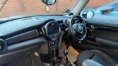The image shows the interior of a modern mini car with a black dashboard, round center display, manual gear stick, and plaid-patterned seats. The steering wheel is on the right, and sunlight and condensation are visible on the windows.