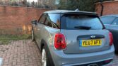 A grey Mini with the license plate YE67 AFX is parked on a brick driveway beside a wall and another car. The weather is clear, and the surroundings include some grass, bushes, and a house in the background—perfect for any mini trader.