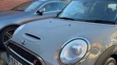 A gray mini car with condensation on the hood is parked beside a silver car in a driveway. The Mini, with license plate YE67 AFX, stands out against brick houses and a clear blue sky in the background.
