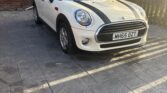 A white Mini Cooper, the classic mini car with black racing stripes, is parked on a paved driveway in front of a brick house. The car's registration plate reads "MH65 DZT." Sunlight casts long shadows near two windows and a door.
