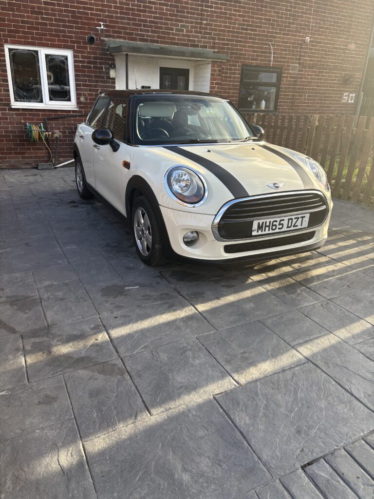 A white Mini Cooper, the classic mini car with black racing stripes, is parked on a paved driveway in front of a brick house. The car's registration plate reads "MH65 DZT." Sunlight casts long shadows near two windows and a door.