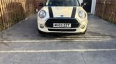 A white Mini car with black racing stripes is parked on a paved driveway in front of a brick house with a black door and wooden fence. The license plate reads “MH65 DZT.” A leafless tree stands to the left—a great find for any Mini trader.