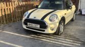 A cream Mini Cooper, a classic mini car with black racing stripes, is parked on a gray paved driveway in front of a brick house with white-framed windows. Bare tree branches and sunlight cast shadows beside the wooden fence.