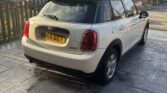 A classic white mini car with a black roof and UK plate "MH65 DZT" is parked on a stone driveway by a wooden fence. Brick houses and a cloudy sky form the backdrop, as the mini is seen from the rear left side.