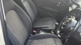 A photo of a mini car’s front interior shows two black fabric seats, a manual gear stick, cup holders, and a dashboard with round air vents. The car door is open, revealing a wooden fence and greenery outside the window.