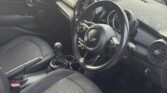 Interior of a right-hand drive mini car, featuring black fabric seats, a manual gear shift, and a steering wheel with the Mini logo. Circular air vents and dials adorn the dashboard. Sunlight highlights the seats, with a fence and bins visible through the window.
