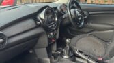 The photo shows the interior of a right-hand drive Mini car. The dashboard has a round central display, manual gear stick, and black fabric seats. The steering wheel is on the right, with a wooden fence and parked red car visible outside.