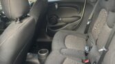 The image shows the backseat interior of a mini car. It features dark fabric seats, visible seatbelts, and cup holders in the center. The door has a round speaker and handle. Sunlight reveals a wooden fence and garden outside the window.